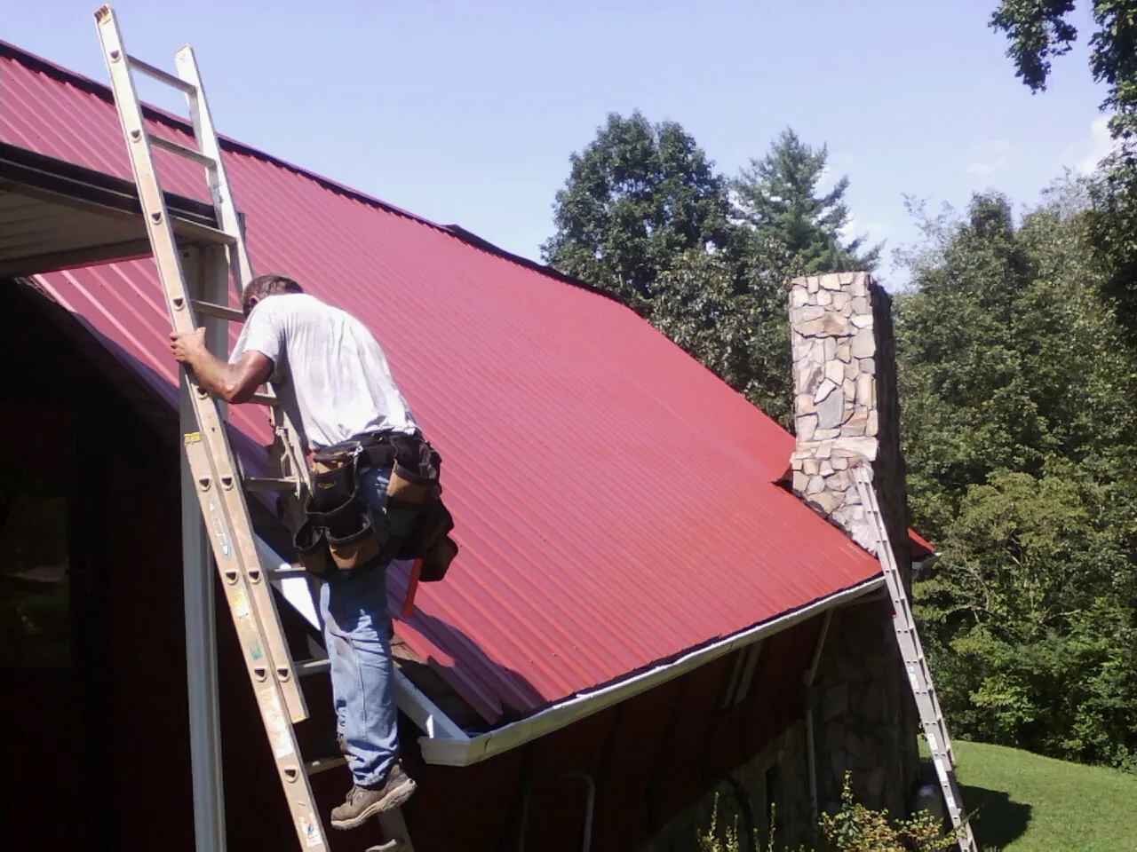 Roofer on a ladder beside a red metal roof and stone chimney — Soffit & Fascia Repair by Roofing Brothers in Kirtland Afb