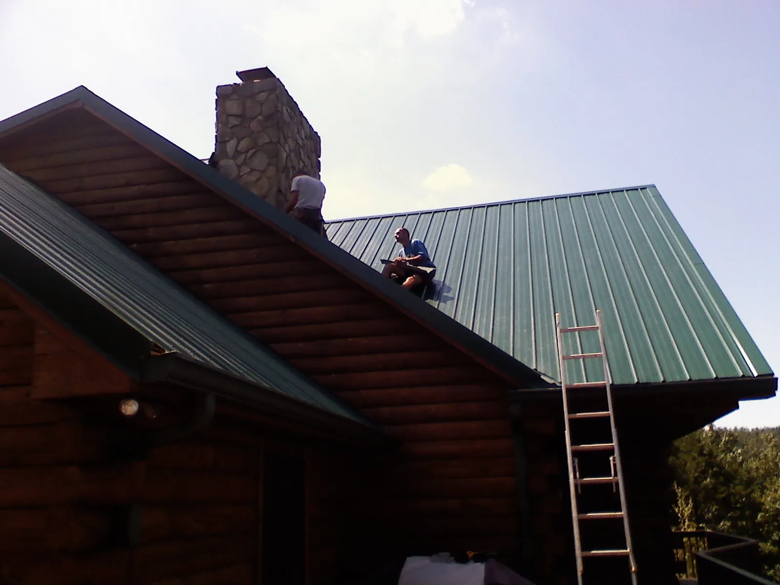 Roofers installing green metal panels beside a stone chimney on a log home in Kirtland Afb